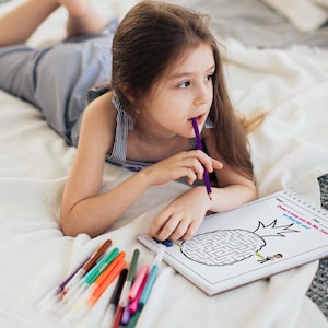 May include: A young girl lies on a white bedspread, concentrating on a black and white maze puzzle printed on a white sheet of paper. The puzzle features a pineapple with the text "Draw a way out for Mr. Tom so he can eat the core!" The girl is holding a purple pencil in her mouth and has a variety of colored markers beside her.
