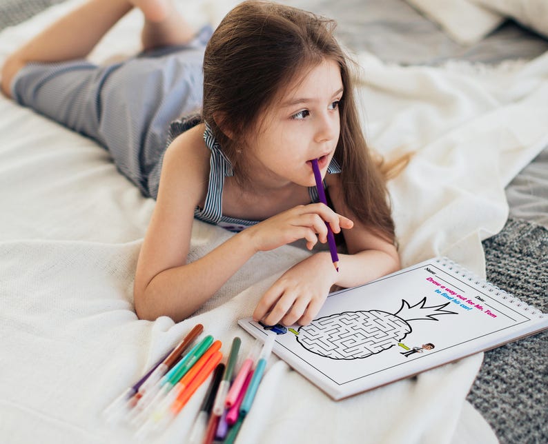 May include: A young girl lies on a white bedspread, concentrating on a black and white maze puzzle printed on a white sheet of paper. The puzzle features a pineapple with the text "Draw a way out for Mr. Tom so he can eat the core!" The girl is holding a purple pencil in her mouth and has a variety of colored markers beside her.