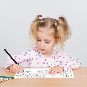 May include: A young girl with blonde pigtails sits at a table, coloring in a maze with a black pencil. The maze is printed on a white sheet of paper with green accents. The girl is wearing a pink and white polka dot shirt.