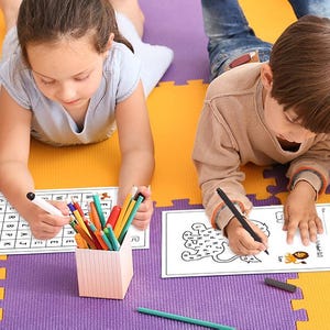 May include: Two children are working on activity sheets. One child is working on a word search puzzle with colorful pencils and a black marker. The other child is working on a maze puzzle with a black marker. Both activity sheets are on a purple and yellow play mat.