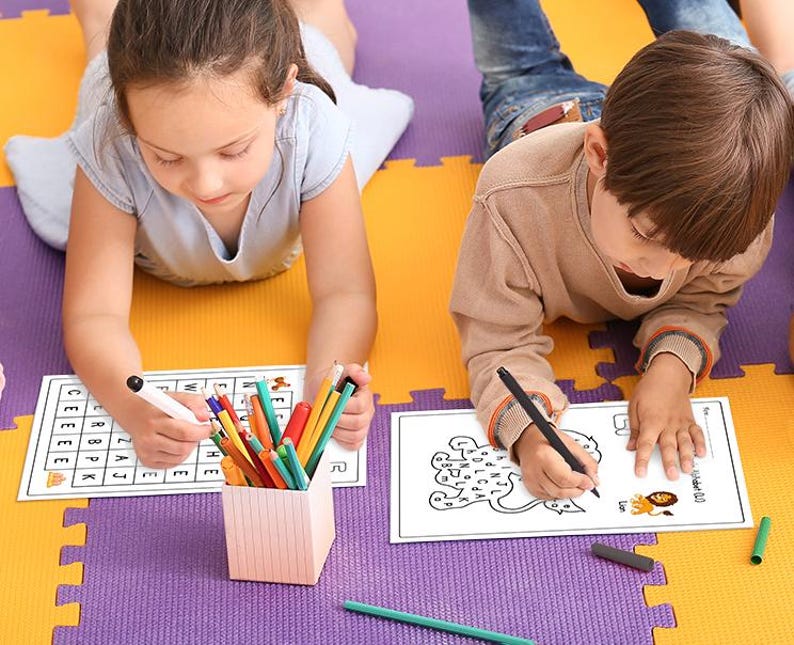 May include: Two children are working on activity sheets. One child is working on a word search puzzle with colorful pencils and a black marker. The other child is working on a maze puzzle with a black marker. Both activity sheets are on a purple and yellow play mat.