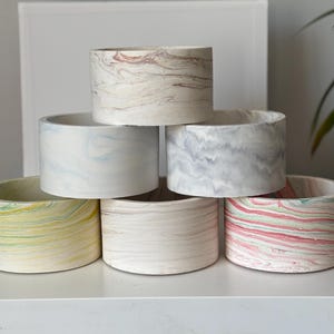 May include: A stack of six cylindrical planters with a marbled design. The planters feature a variety of colors, including white, gray, pink, yellow, and blue. The planters are arranged on a white surface.