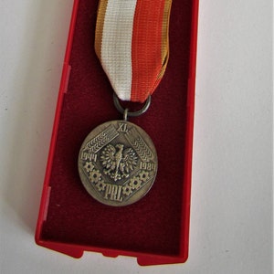 Puede incluir: Medalla de plata con cinta roja, blanca y dorada, presentada en un estuche rojo. La medalla presenta un emblema de águila y el texto "1944-1984". El estuche está abierto, revelando la medalla y la cinta.