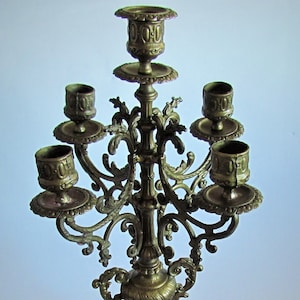 Può includere: Un candelabro in ottone a cinque bracci con dettagli decorativi e una base a rotolo. Il candelabro è in metallo dorato e ha uno stile vintage.