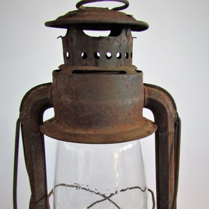 Könnte beinhalten: Eine antike, rostige Öllampe aus Metall mit einer klaren Glaskugel und einem Drahtkäfig. Die Lampe hat einen Metallgriff und einen runden Sockel.