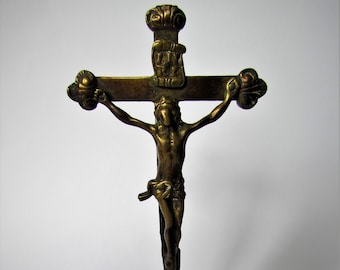 May include: A small, ornate brass crucifix with a detailed figure of Jesus Christ on the cross. The cross is mounted on a square base with intricate engravings.