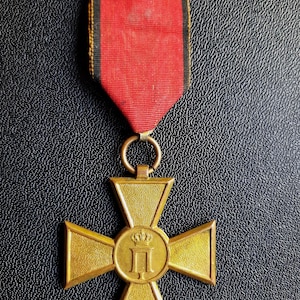 Puede incluir: Una medalla dorada en forma de cruz con una cinta roja. La medalla presenta un emblema circular central con una corona y una letra. La cinta es roja con rayas negras en los bordes.