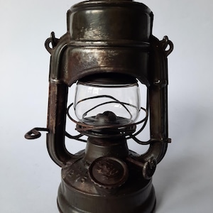 Op de afbeelding: Een donkerbruine, verweerde metalen olielantaarn met een heldere glazen bol beschermd door een draadkooi. De lantaarn heeft een ronde voet, een handvat aan de bovenkant en een zijdelingse draaiknop. Het metalen oppervlak vertoont tekenen van ouderdom.