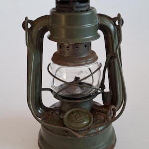 Può includere: Una lanterna da tempesta in metallo verde oliva con un globo in vetro trasparente, una gabbia in filo metallico e una maniglia superiore. La lanterna presenta una base circolare, un tappo del serbatoio laterale e segni di ruggine e usura.