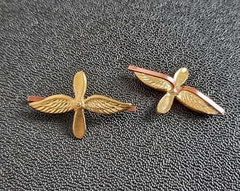 Puede incluir: Dos pines de metal de color dorado en forma de hélice. Un pin es un diseño de hélice simple, mientras que el otro tiene alas a cada lado de la hélice.