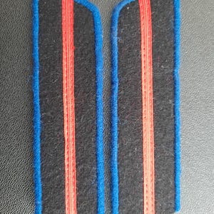Peut inclure: Deux épaulettes en tissu noir avec des bordures rouges et bleues. Les épaulettes ont deux bandes rouges sur chacune.