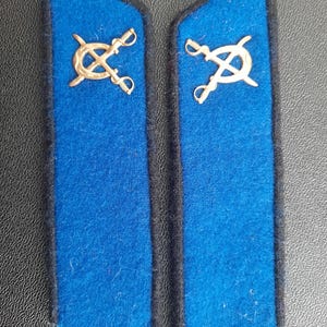 Puede incluir: Dos hombreras azules con espadas cruzadas doradas y un emblema circular.