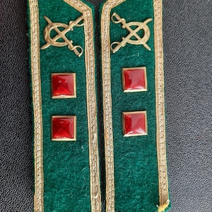 May include: A pair of green military shoulder boards with gold trim and red squares. The shoulder boards have a gold emblem with crossed swords and a crescent moon.