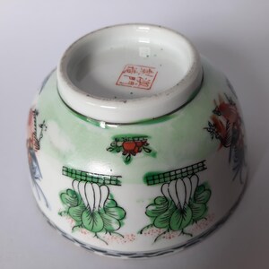 May include: An upside-down, antique porcelain bowl with a detailed floral and bird design. The bowl features a white base with green and red accents, and a red stamp on the bottom. The rim is trimmed with a thin black line.