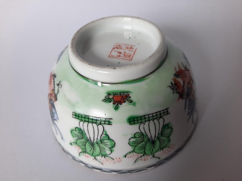 May include: An upside-down, antique porcelain bowl with a detailed floral and bird design. The bowl features a white base with green and red accents, and a red stamp on the bottom. The rim is trimmed with a thin black line.