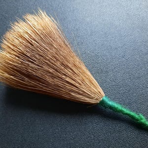 Op de afbeelding: Een kleine, handgemaakte bezem met een waaiervormige kop van lichtbruine borstelharen. De steel is omwikkeld met groen garen. De bezem staat tegen een donkere, getextureerde achtergrond.
