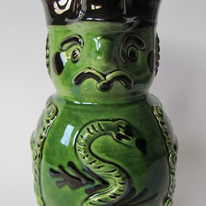 May include: A green ceramic pitcher with a brown hat and a snake design. The pitcher has a whimsical face with a mustache.