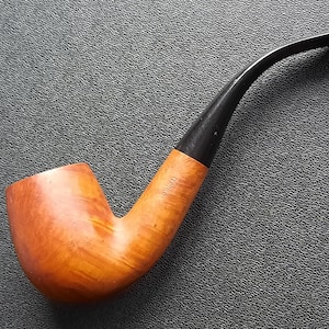 May include: A classic tobacco pipe with a curved stem. The pipe features a light brown bowl and a black stem. The bowl has a natural wood grain finish, and the stem is smooth and curved. The pipe is designed for smoking.