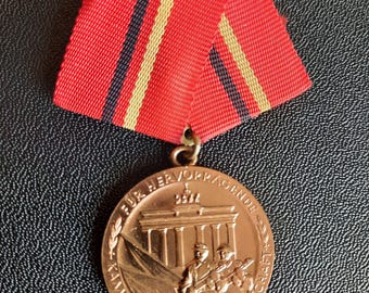 May include: A bronze-colored circular medal featuring an embossed image of the Brandenburg Gate and two soldiers with rifles. The text "FUR HERVORRAGENDE KAMPF-UND EINSATZBEREITSCHAFT" is inscribed along the edge. It hangs from a red ribbon with thin black and yellow stripes.