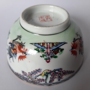 May include: A small, white porcelain bowl with colorful hand-painted designs. The bowl features a light green background and is decorated with birds, flowers, and geometric patterns in red, blue, and other colors. A red stamp is visible on the bottom.