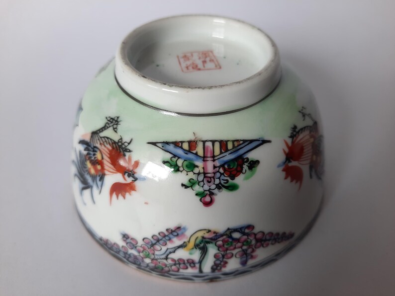 May include: A small, white porcelain bowl with colorful hand-painted designs. The bowl features a light green background and is decorated with birds, flowers, and geometric patterns in red, blue, and other colors. A red stamp is visible on the bottom.