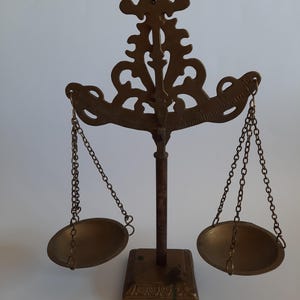 Puede incluir: Una balanza de metal color latón con un poste vertical central con una parte superior decorativa, una base cuadrada y dos platos circulares suspendidos por cadenas de metal delgadas. La barra horizontal muestra marcas numéricas de medición.