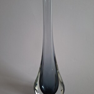 Könnte beinhalten: Eine hohe, schlanke Glasvase mit einer tropfenförmigen Basis, die in einen langen, schmalen Hals übergeht. Die Vase besteht aus klarem Glas mit einem tiefen, rauchig grau-blauen Kern, der sich nach oben hin verjüngt und eine glatte, solide Silhouette bildet.