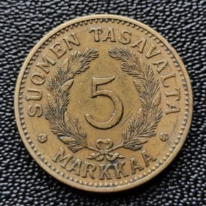 May include: A bronze Finnish coin with the text "Suomen Tasavalta" and "5 Markkaa" around a wreath of pine branches. The number "5" is in the center of the coin.