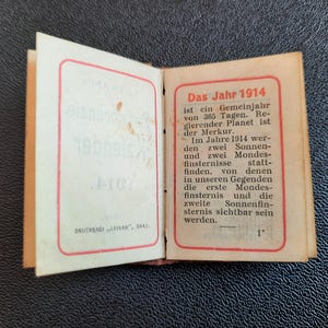 Puede incluir: Un peque&ntilde;o libro abierto con una cubierta roja y blanca. El texto de la cubierta dice "Das Jahr 1914" y describe el a&ntilde;o como un a&ntilde;o com&uacute;n de 365 d&iacute;as. El texto tambi&eacute;n menciona que Mercurio es el planeta regente y que habr&aacute; dos eclipses solares y dos eclipses lunares en 1914.