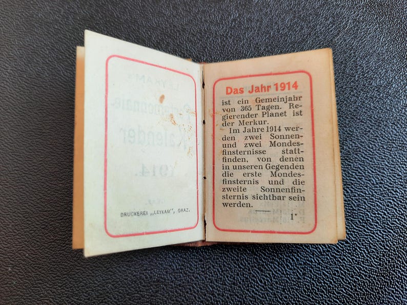 Puede incluir: Un peque&ntilde;o libro abierto con una cubierta roja y blanca. El texto de la cubierta dice "Das Jahr 1914" y describe el a&ntilde;o como un a&ntilde;o com&uacute;n de 365 d&iacute;as. El texto tambi&eacute;n menciona que Mercurio es el planeta regente y que habr&aacute; dos eclipses solares y dos eclipses lunares en 1914.