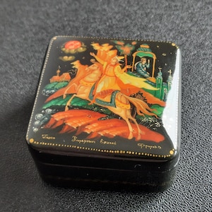 Può includere: Scatola laccata nera con una scena dipinta a mano raffigurante una figura a cavallo, una figura seduta ed elementi architettonici. L'opera d'arte è in colori vivaci, con accenti dorati e testo in basso.