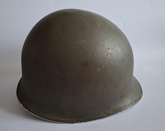 May include: A dome-shaped olive drab military helmet with a smooth, rounded crown and a slightly flared brim. The surface of the helmet shows signs of wear, including minor surface scuffs and small dark spots across the exterior shell.