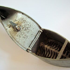 May include: A vintage cast iron flat iron with a hinged lid and a grate inside. The iron is silver in color and has a worn, aged appearance.