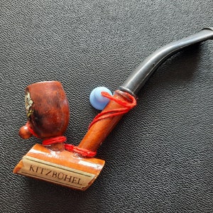 May include: A wooden pipe with a red string wrapped around it. The bowl of the pipe is decorated with a floral design and the word "KITZBÜHEL" is written on the side.
