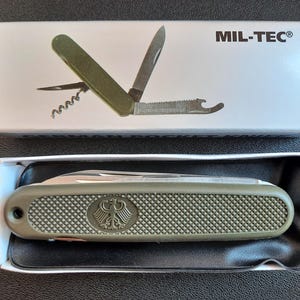 May include: A green multi-tool pocket knife with a black handle and a silver blade. The knife has a bottle opener, a corkscrew, and a saw. The knife is in a white box with the words "MIL-TEC" printed on it.