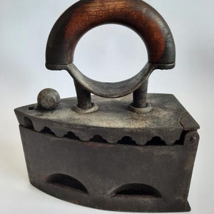 Op de afbeelding: Een vintage gietijzeren houtskoolstrijkijzer met een houten handvat. Het strijkijzer heeft een afgeronde basis met een reeks gaten voor luchtcirculatie. Het handvat is met een metalen ring aan het strijkijzer bevestigd.
