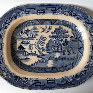 Könnte beinhalten: Eine ovale blaue und weiße Porzellan-Servierplatte mit einem Weidenmuster. Die Platte hat einen erhöhten Rand und einen gewellten Rand.