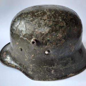 Può includere: Un elmetto militare antico, verde oliva, con una superficie strutturata e usurata. L'elmetto ha una calotta arrotondata, un bordo svasato e diversi dettagli di rivetti. La vernice è scheggiata e usurata, rivelando il metallo sottostante. (circa 20 cm)