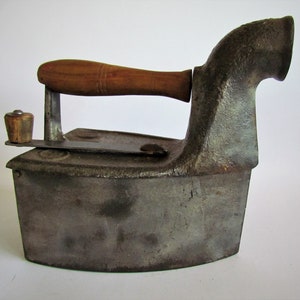 May include: A vintage cast iron flat iron with a wooden handle and a spout for adding fuel. The iron has a gray finish and shows signs of wear and tear.