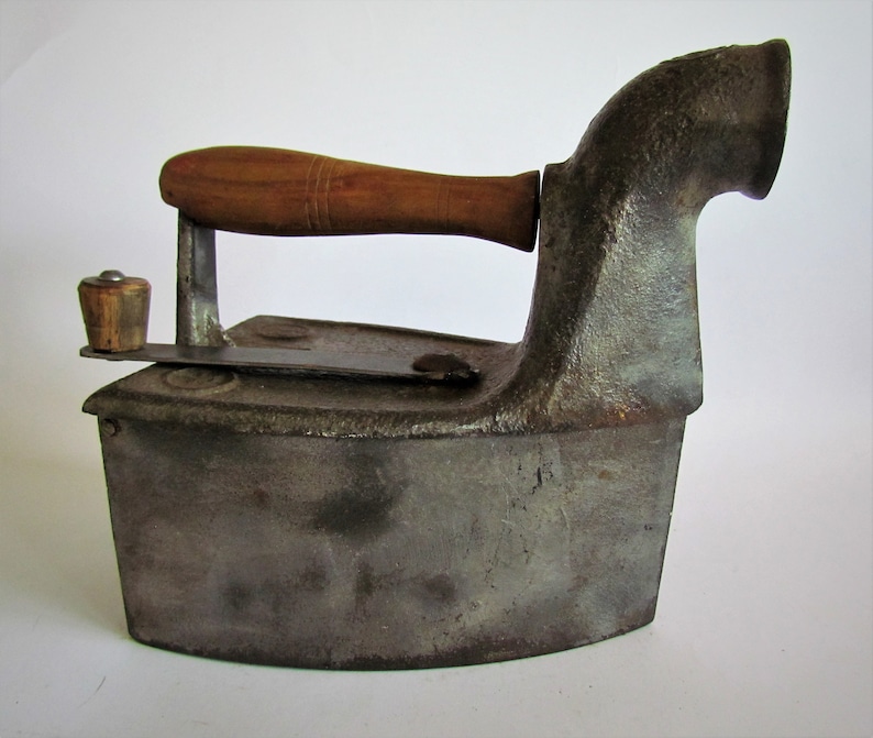 May include: A vintage cast iron flat iron with a wooden handle and a spout for adding fuel. The iron has a gray finish and shows signs of wear and tear.