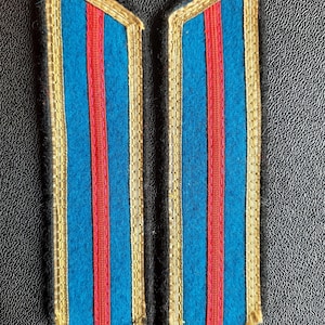May include: Two blue shoulder boards with red and gold trim. The boards are made of fabric and have a traditional military style.