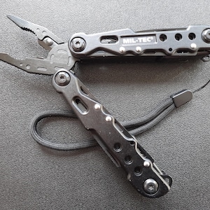 Pode incluir: Ferramenta multifuncional preta com um cordão. A ferramenta tem uma variedade de funções, incluindo alicates, uma faca, uma chave de fenda e um abridor de garrafas. A ferramenta é etiquetada como "MIL-TEC".