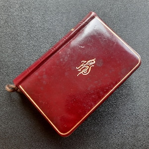 May include: A small, rectangular, burgundy-colored book with gold trim and a gold monogram on the cover. The book has a worn appearance and a brown cord attached to the spine.