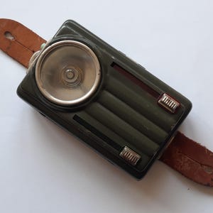 Puede incluir: Una linterna vintage verde oliva con una luz circular y una correa de cuero marrón. La linterna tiene un cuerpo rectangular con detalles acanalados y dos interruptores plateados.