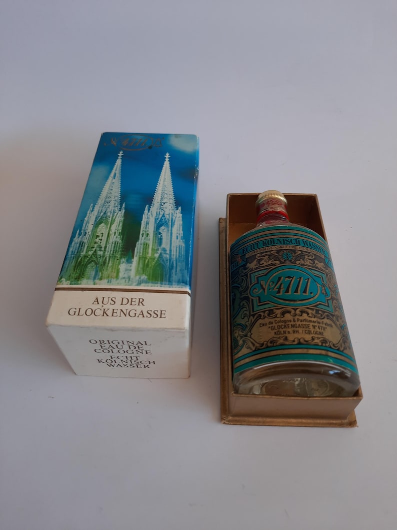 Puede incluir: Una botella de colonia vintage y su caja. La botella es transparente con una etiqueta turquesa y dorada. La caja presenta una ilustraci&oacute;n azul y blanca de una catedral. El texto en la caja dice "AUS DER GLOCKENGASSE" y "ORIGINAL EAU DE COLOGNE".