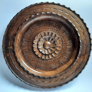 May include: A circular dark brown wooden plate featuring intricate hand-carved geometric patterns and a central floral motif. The rim of the plate has a scalloped edge, and the surface displays a polished finish with visible wood grain textures.