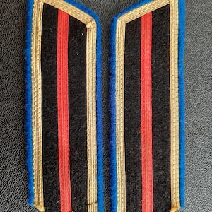 Peut inclure: Deux épaulettes en tissu noir avec des bordures dorées et bleues et une bande rouge au centre.