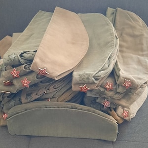 Könnte beinhalten: Ein Stapel olivgrüner Militärkappen mit roten Sternansteckern. Die Kappen sind aus weichem Stoff gefertigt.