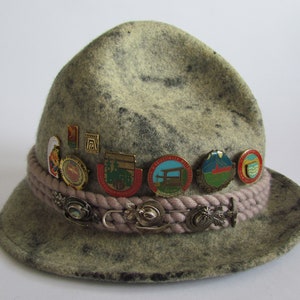 Op de afbeelding: Een groene wollen hoed met een bruin gevlochten band en verschillende kleine spelden. De spelden hebben verschillende ontwerpen, waaronder een bergketen, een brug en een gebouw.