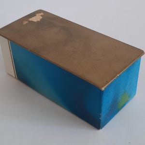 Puede incluir: Caja rectangular con un dise&ntilde;o degradado azul en los lados y una tapa marr&oacute;n. La caja tiene una franja blanca en un lado. La tapa muestra signos de desgaste. La caja es probablemente vintage o antigua.